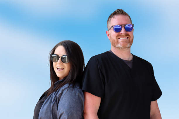A woman and a man stand in front of a blue background smiling and wearing reflective sunglasses.